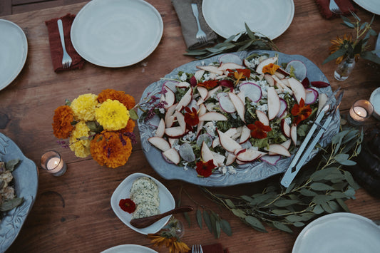 Silver platter of farm to table food with tealight candles, marigolds, herbed butter, foliage, and plates, on a wooden table